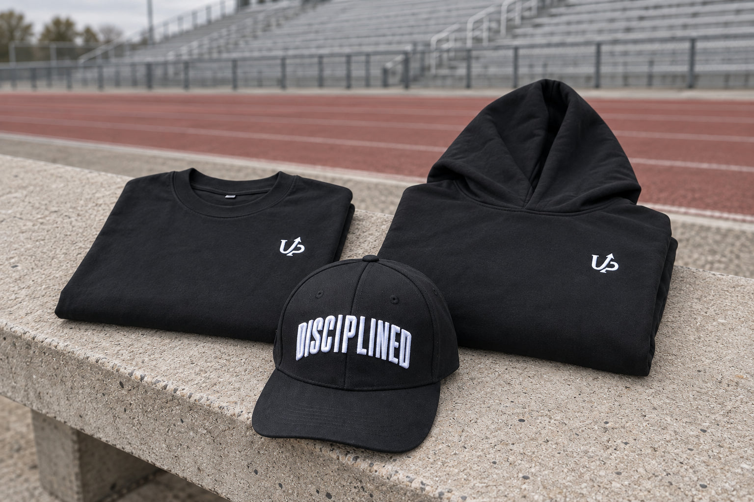 Drop 001 hoodie and tee folded beside the structured hat at an empty running track